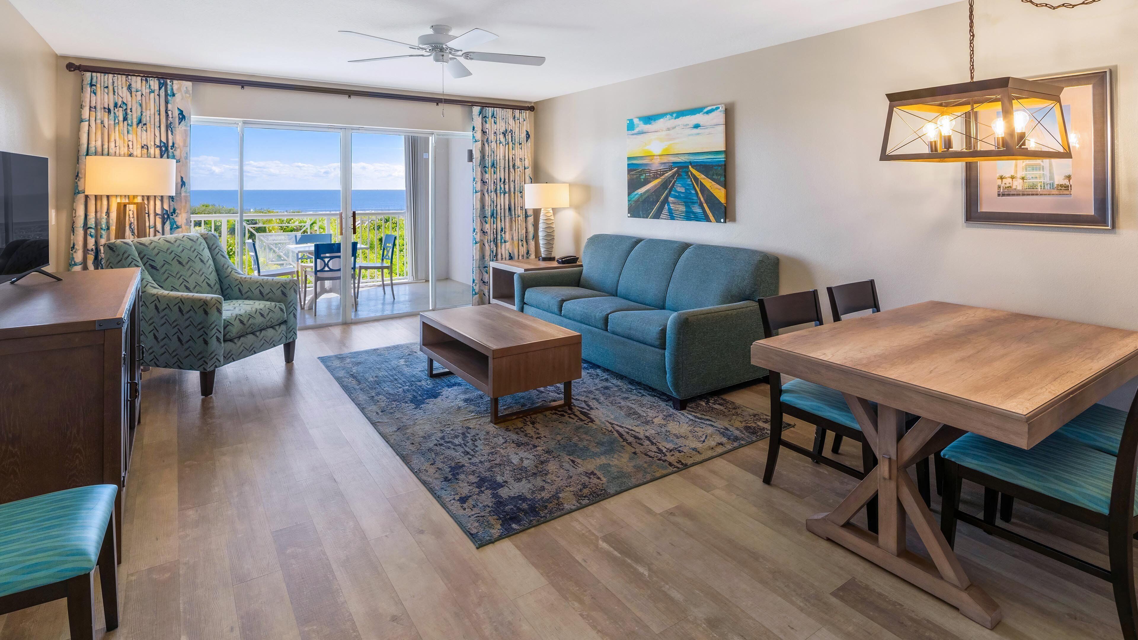 A modern living room with a sofa, coffee table, and dining table, a balcony with a view, and a ceiling fan.