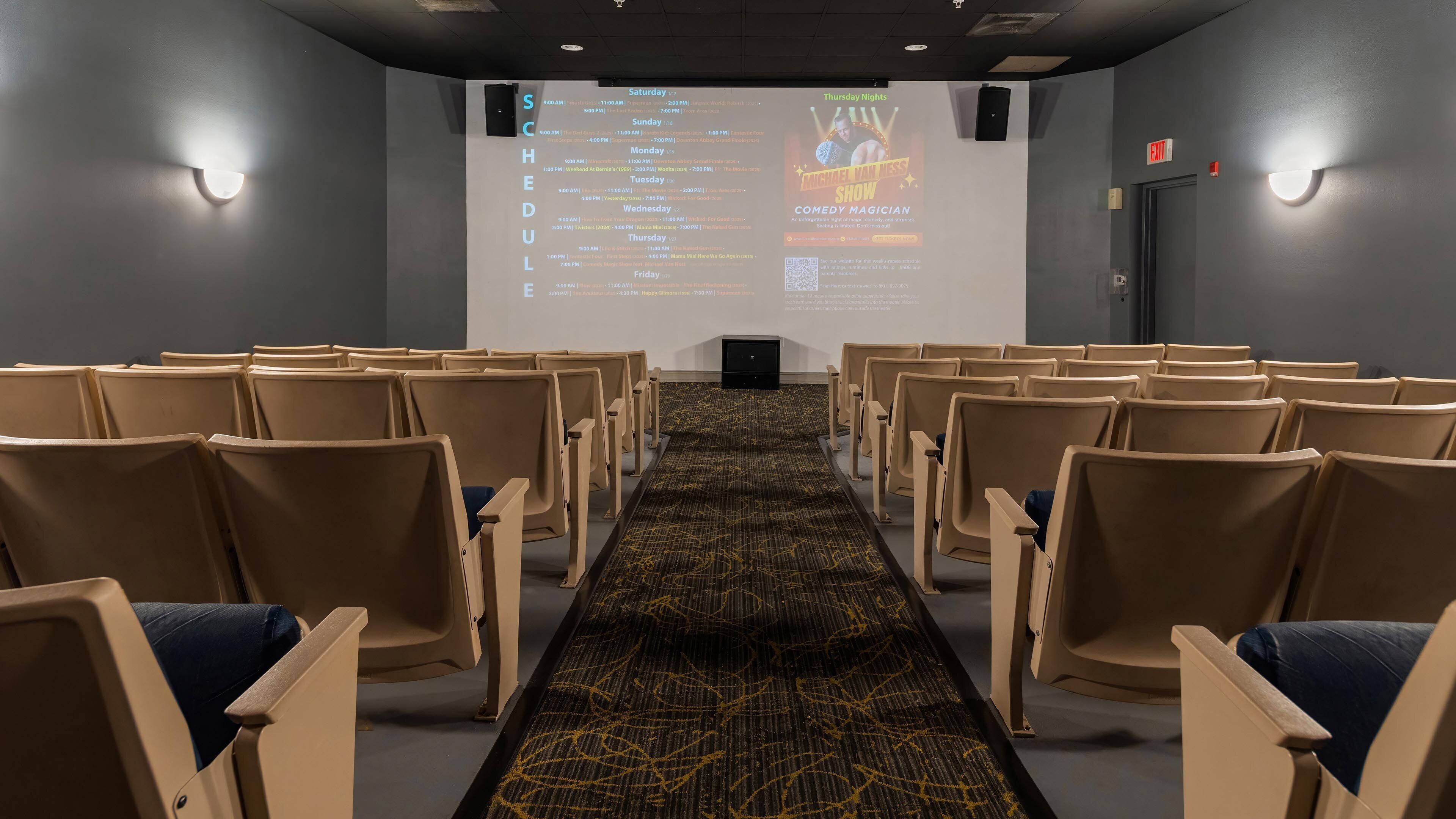 A theater with rows of seats, a carpeted floor, and a screen displaying a schedule.