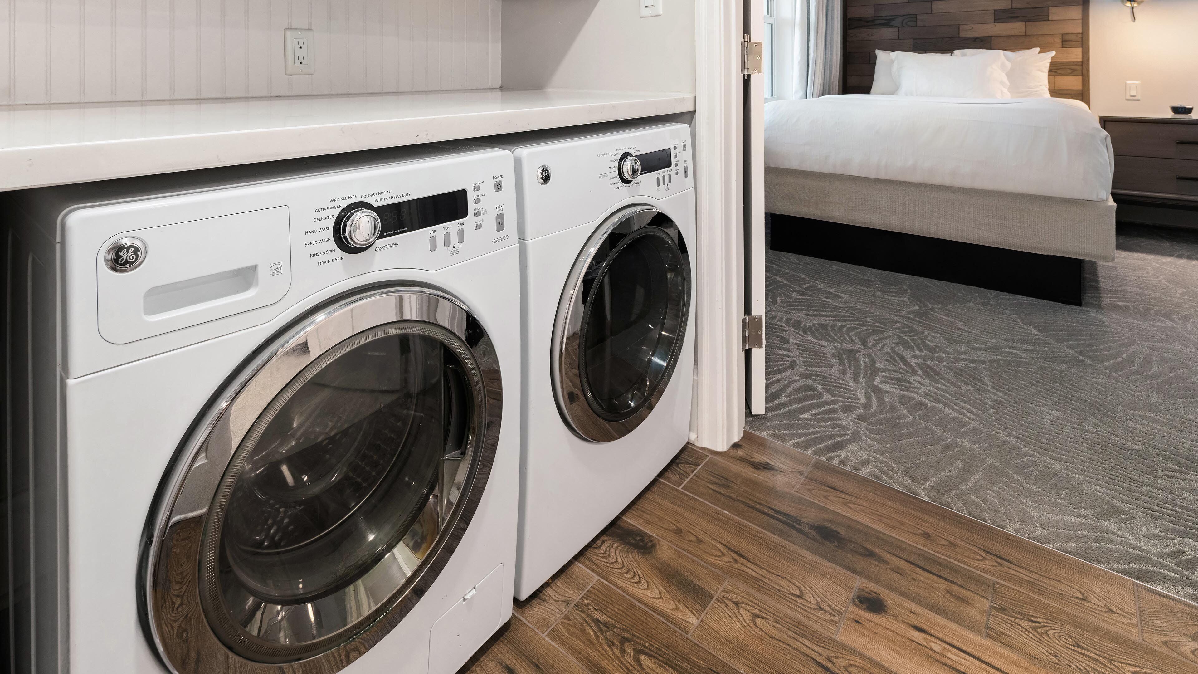Un moderno lavadero con dos lavadoras de carga frontal, una cama con ropa de cama blanca y un cabecero de madera.