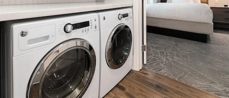 Un moderno lavadero con dos lavadoras de carga frontal, una cama con ropa de cama blanca y un cabecero de madera.