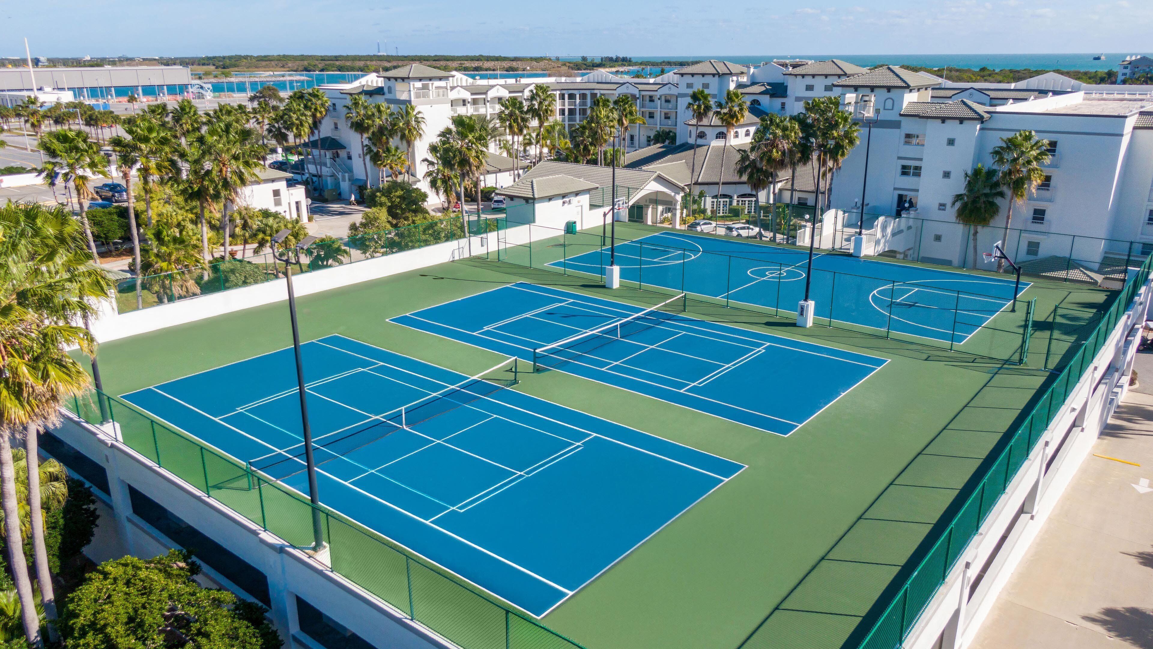 Vue aérienne d’un complexe de courts de tennis comprenant plusieurs terrains, entouré de bâtiments et de palmiers.