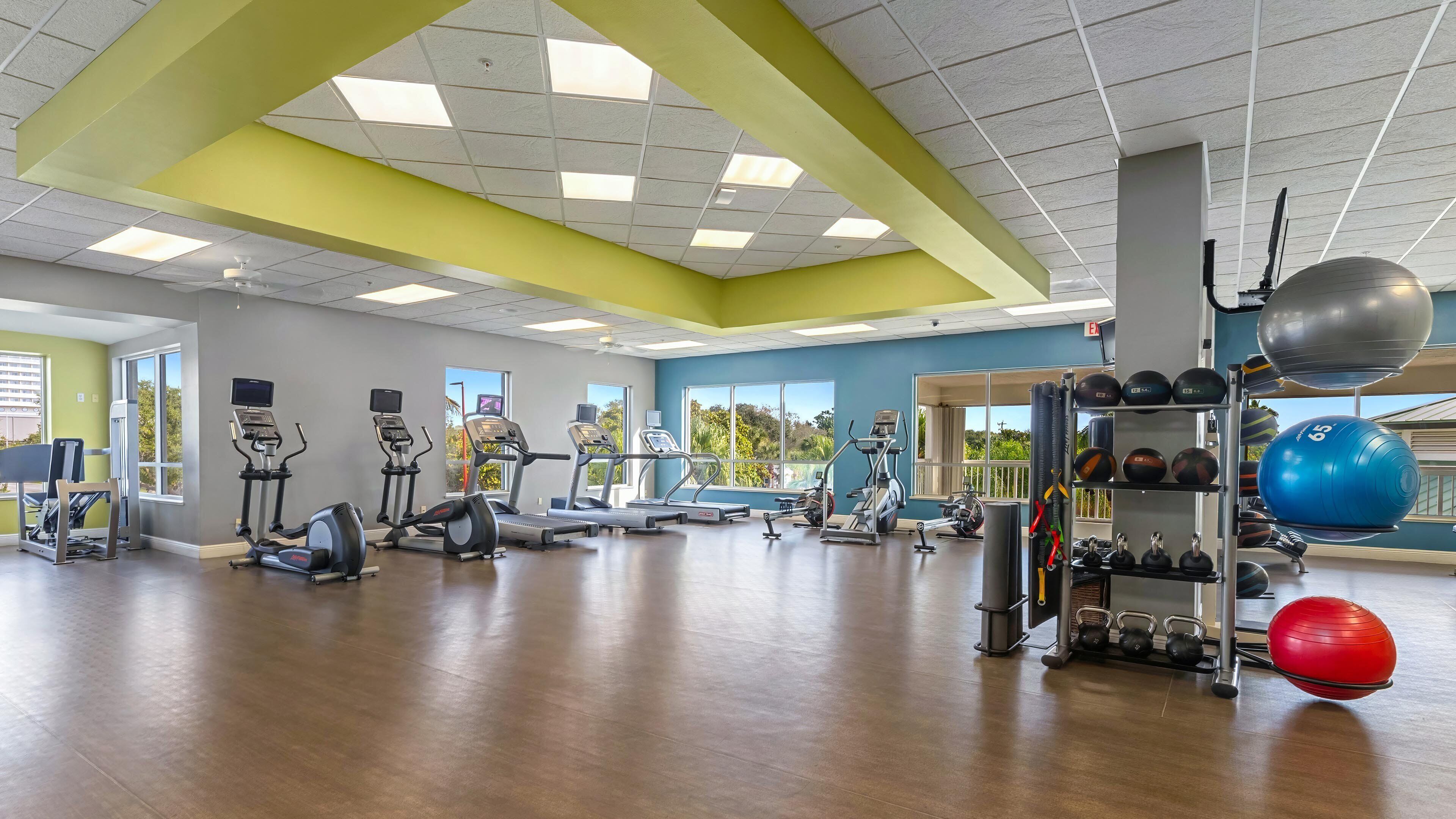A gym with various exercise equipment, large windows, and a view of greenery outside.