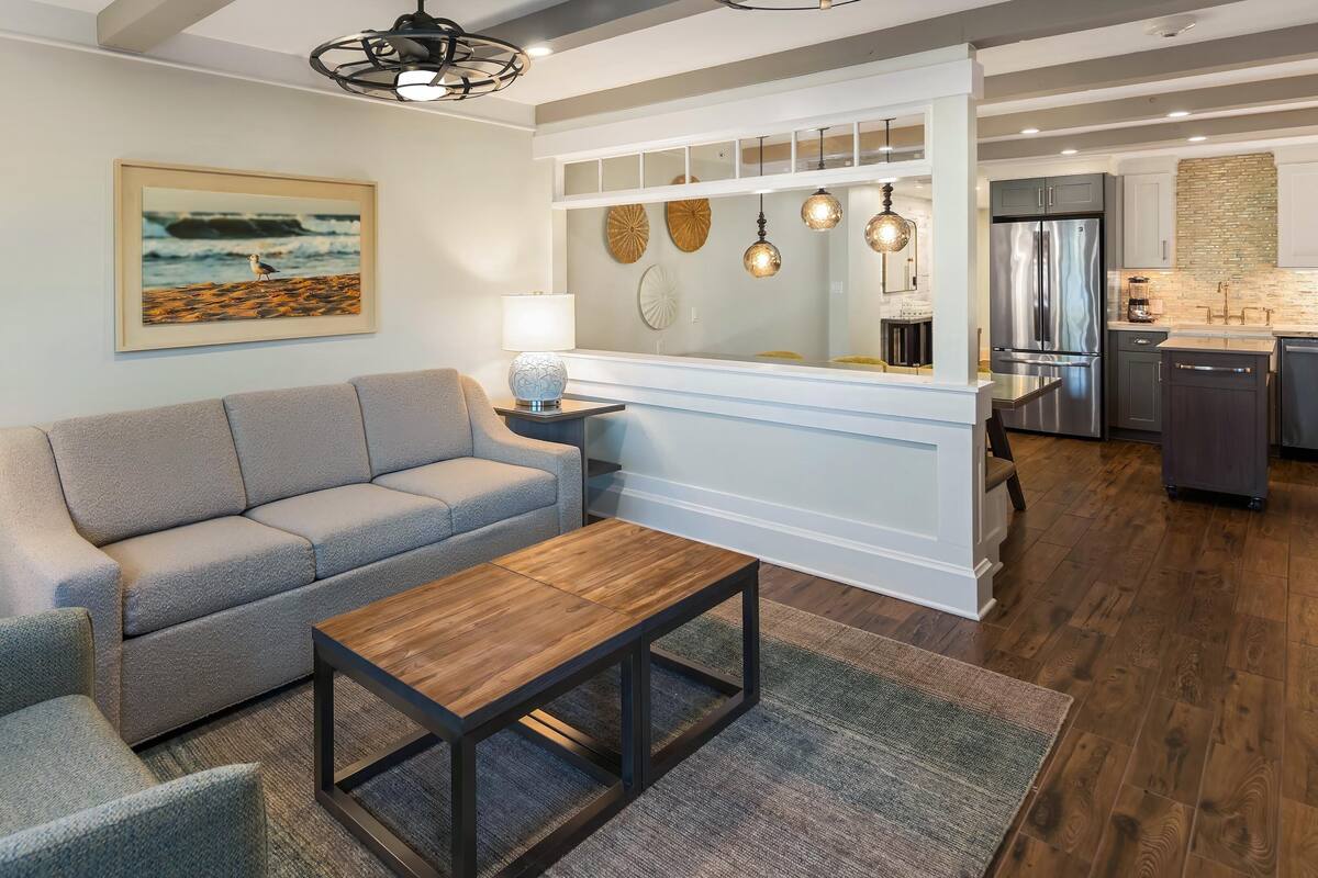 A modern living room with a grey sofa, a wooden coffee table, and a kitchen area with a stainless steel refrigerator and a brick backsplash.