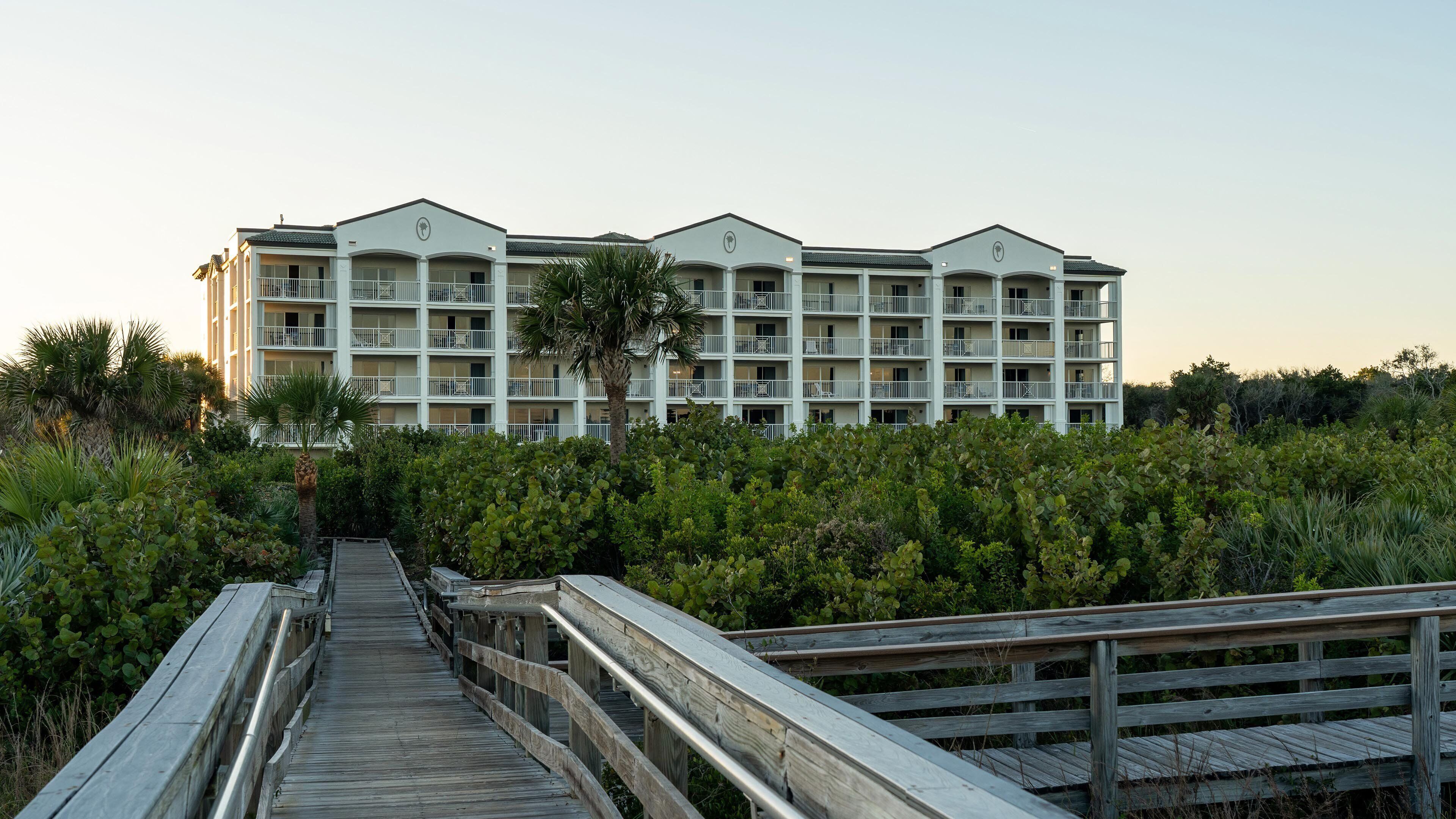 A multi-story hotel with balconies, surrounded by dense vegetation and a wooden boardwalk leading to the entrance.