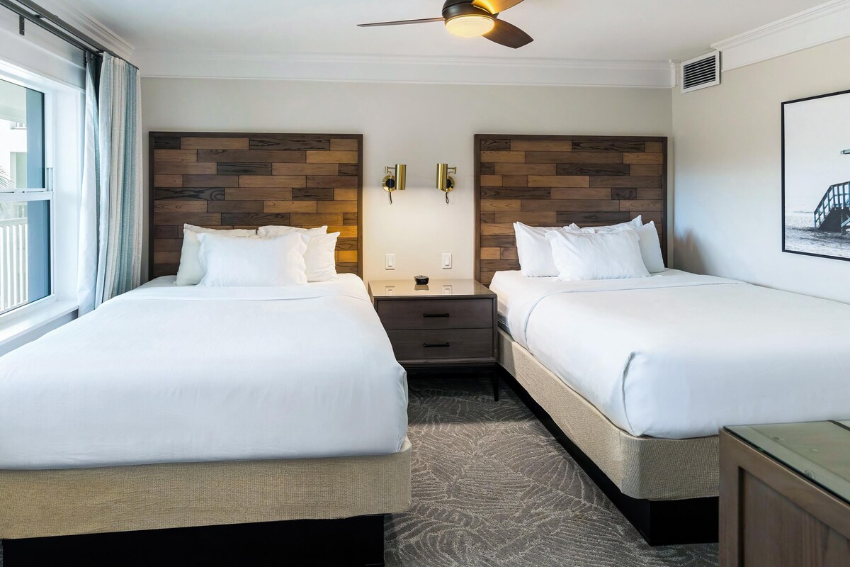 A hotel room with two beds, a ceiling fan, a nightstand, and a window with a view of a balcony.