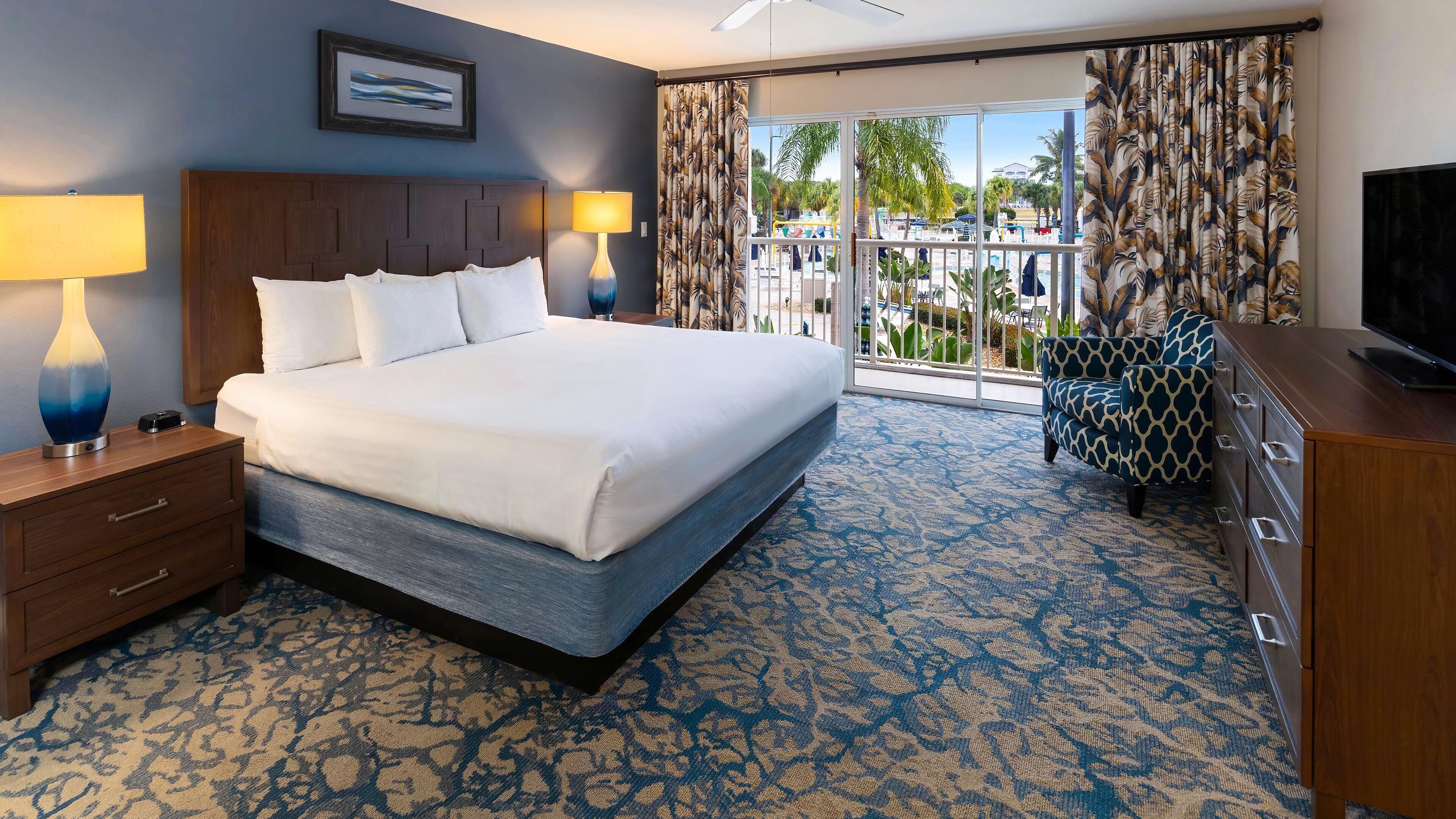 A hotel room with a bed, nightstands, a TV, and a view of a pool area.