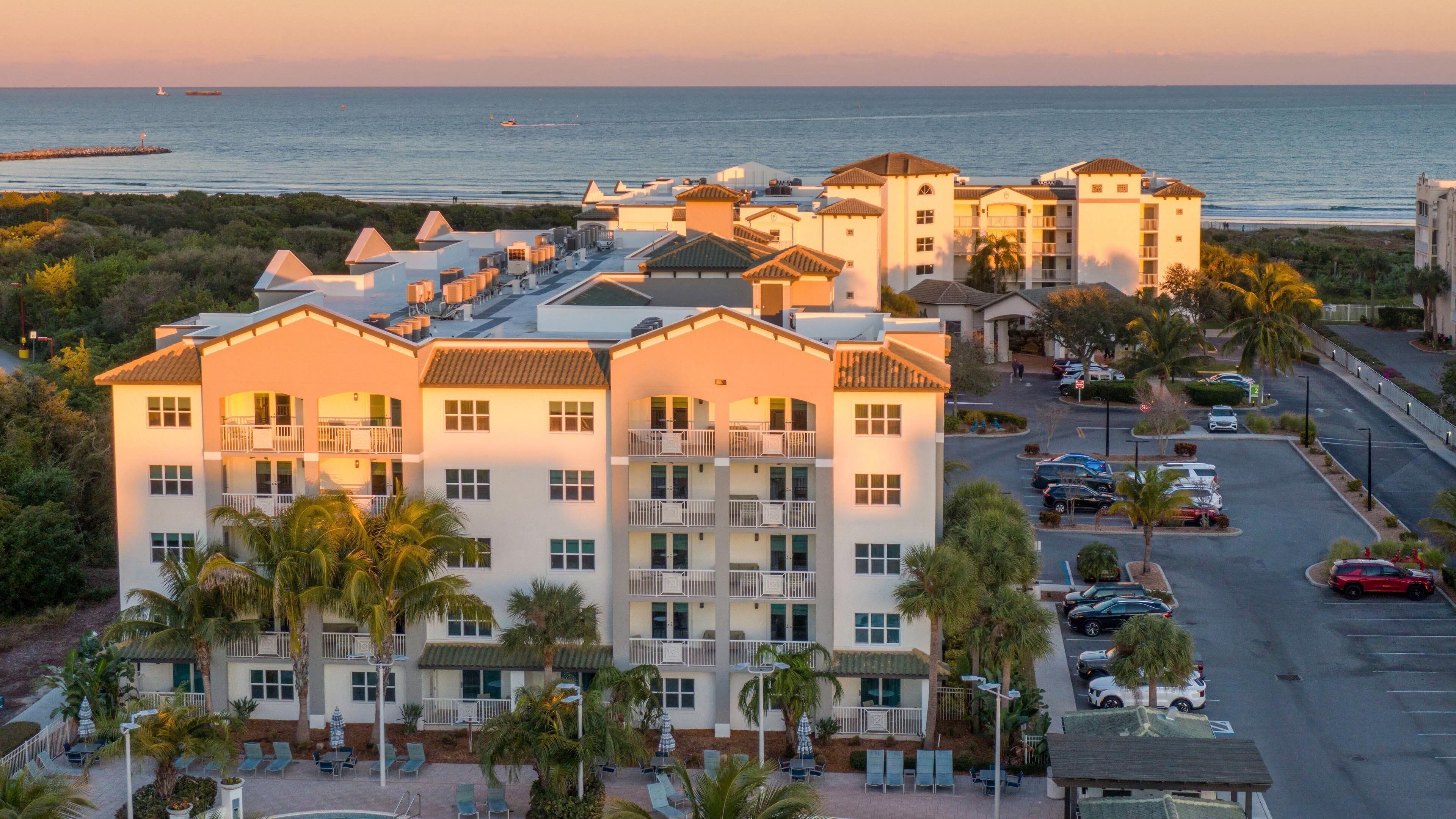 A multi-story hotel with balconies, a parking lot, and a view of the ocean.