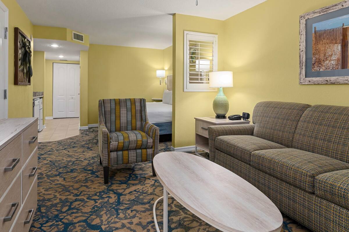 A hotel room with a bed, a sofa, a chair, a small table, a TV, and a painting on the wall.