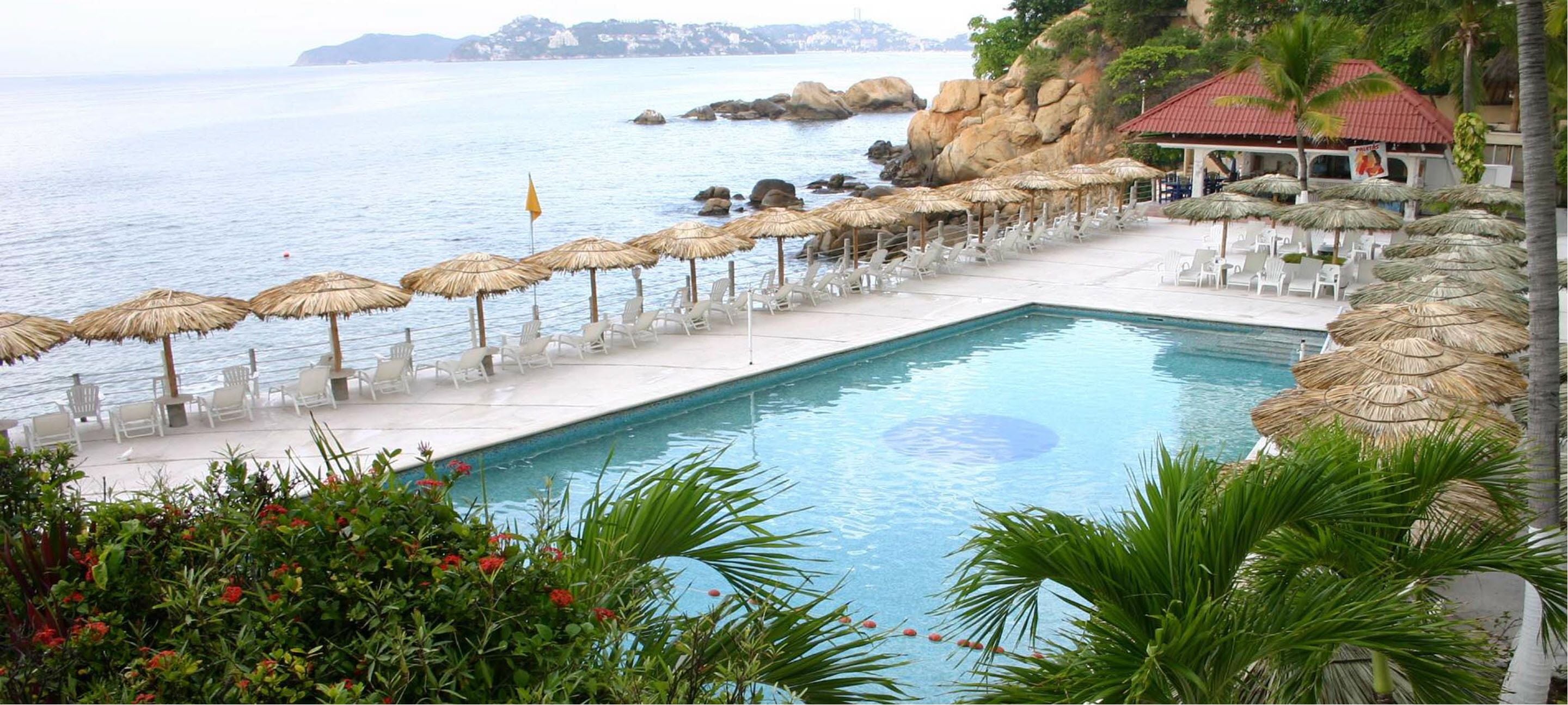 A pool with lounge chairs and umbrellas, a view of the sea, and a building with a red roof.