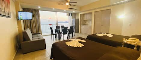A hotel room with two beds, a sofa, a TV, a dining area with chairs, and a view of the sea.