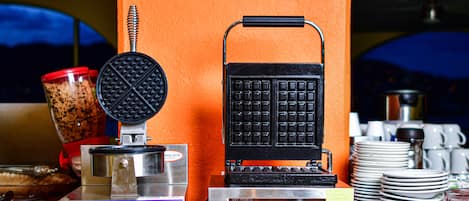 A waffle iron, a coffee maker, and a blender on a counter with plates and cups stacked nearby.
