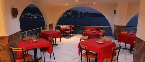 A restaurant with red tablecloths and chairs, a ceiling fan, and a view of a city at night.