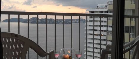 A balcony with a table set for two, overlooking a body of water and a distant shoreline.