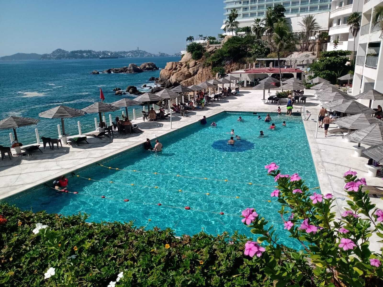 Uma área de piscina com águas cristalinas, rodeada por espreguiçadeiras e guarda-sóis, com vista para a costa, edifícios e palmeiras.