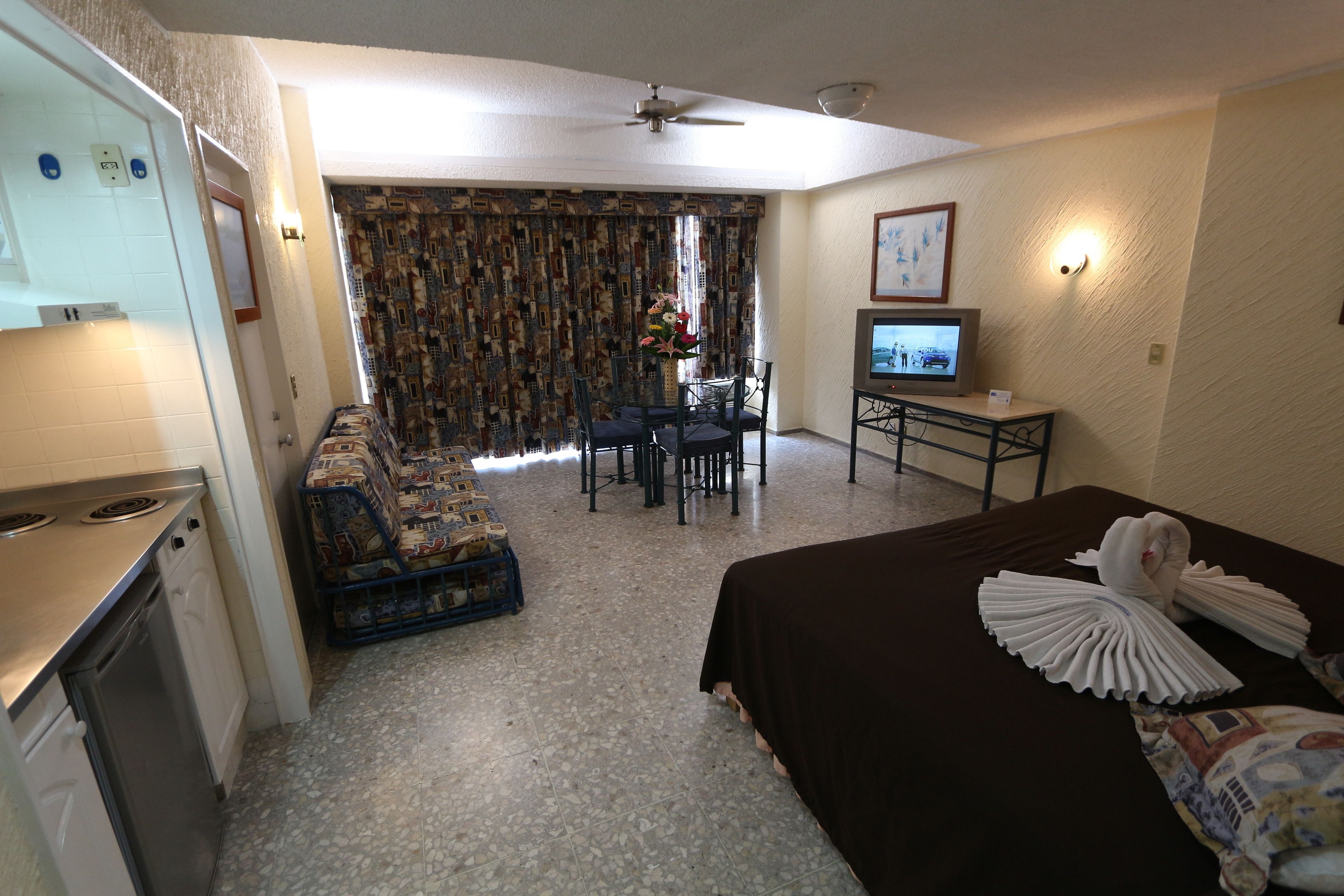 Una habitación de hotel con kitchenette, un comedor con mesa y sillas, una cama con almohadas decorativas y un televisor sobre una mesita.