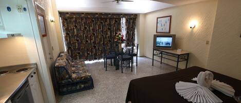 Una habitación de hotel con kitchenette, un comedor con mesa y sillas, una cama con almohadas decorativas y un televisor sobre una mesita.