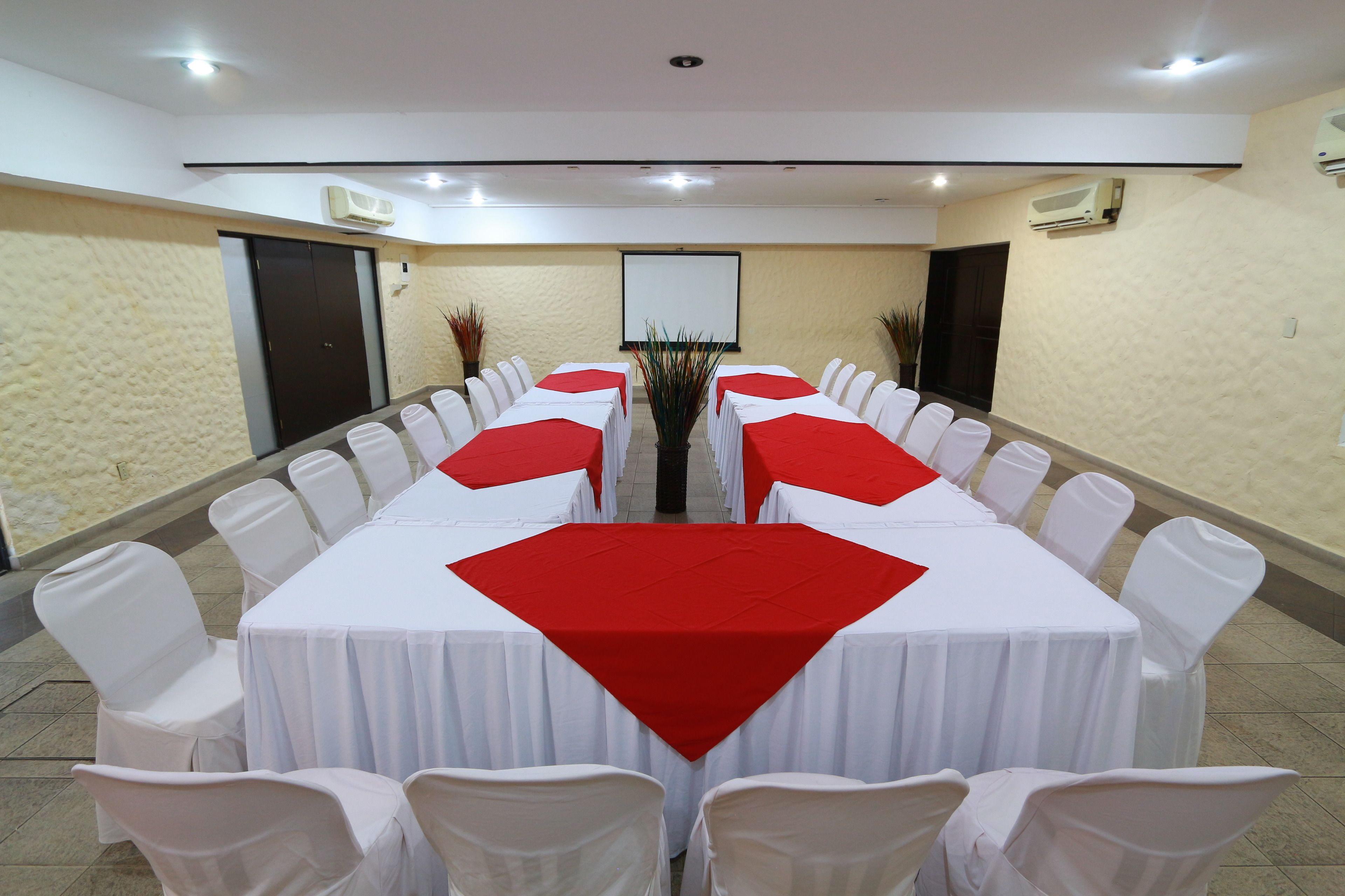 Uma sala de conferências com uma mesa comprida, cadeiras brancas e jogos americanos vermelhos.