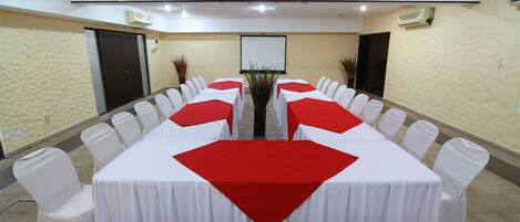 Uma sala de conferências com uma mesa comprida, cadeiras brancas e jogos americanos vermelhos.