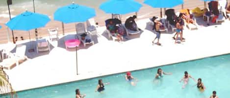 Un área de piscina con múltiples sombrillas azules, sillas de descanso y gente nadando y relajándose.