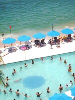 Área de piscina com vários guarda-sóis azuis, espreguiçadeiras e pessoas nadando e relaxando.