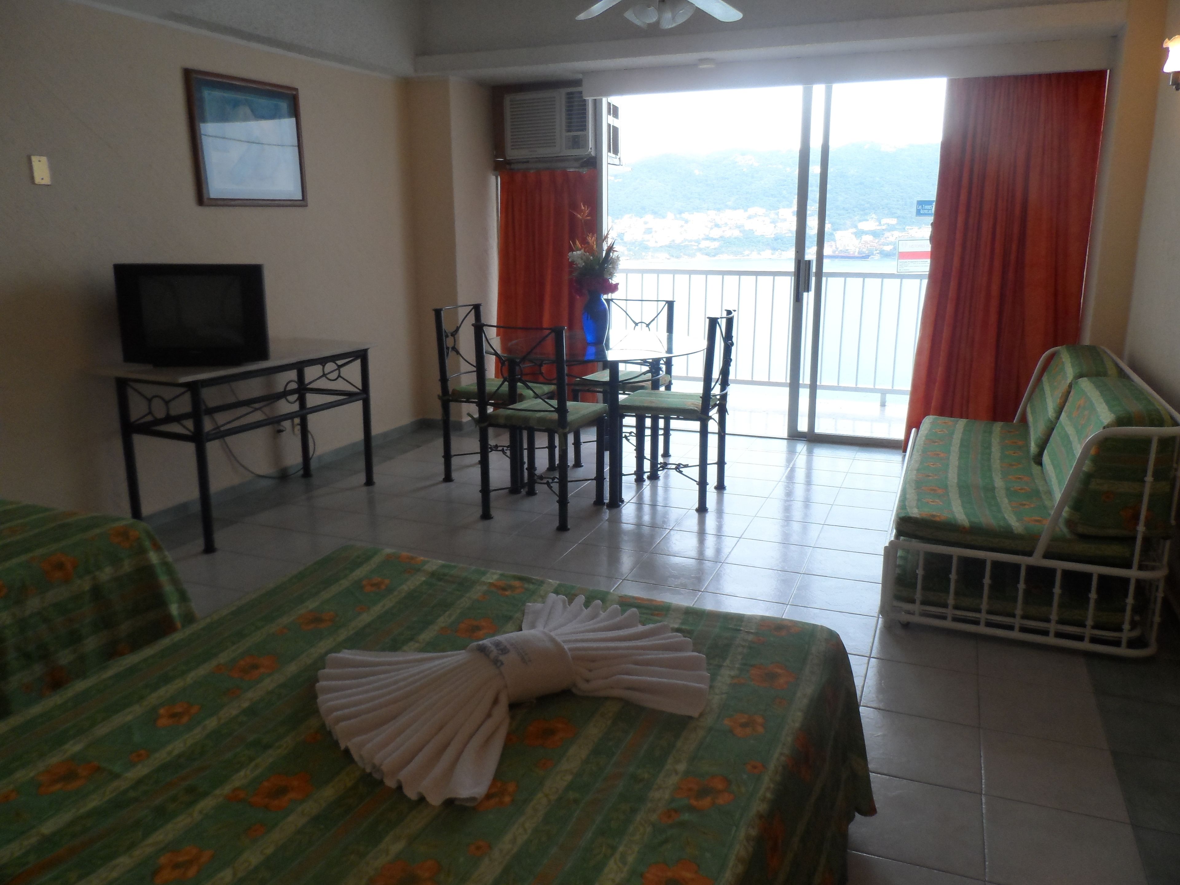 A hotel room with two beds, a TV, a table with chairs, and a balcony with a view of the city.