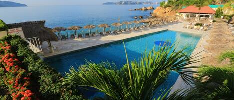 A pool area with lounge chairs and umbrellas, a view of the sea, and a building with a red roof.