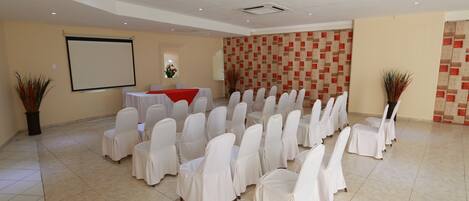 Un salón de conferencias con filas de sillas cubiertas con manteles blancos, una pantalla de proyector y una pequeña mesa con un paño rojo.