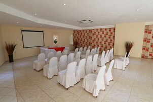 Un salón de conferencias con filas de sillas cubiertas con manteles blancos, una pantalla de proyector y una pequeña mesa con un paño rojo.