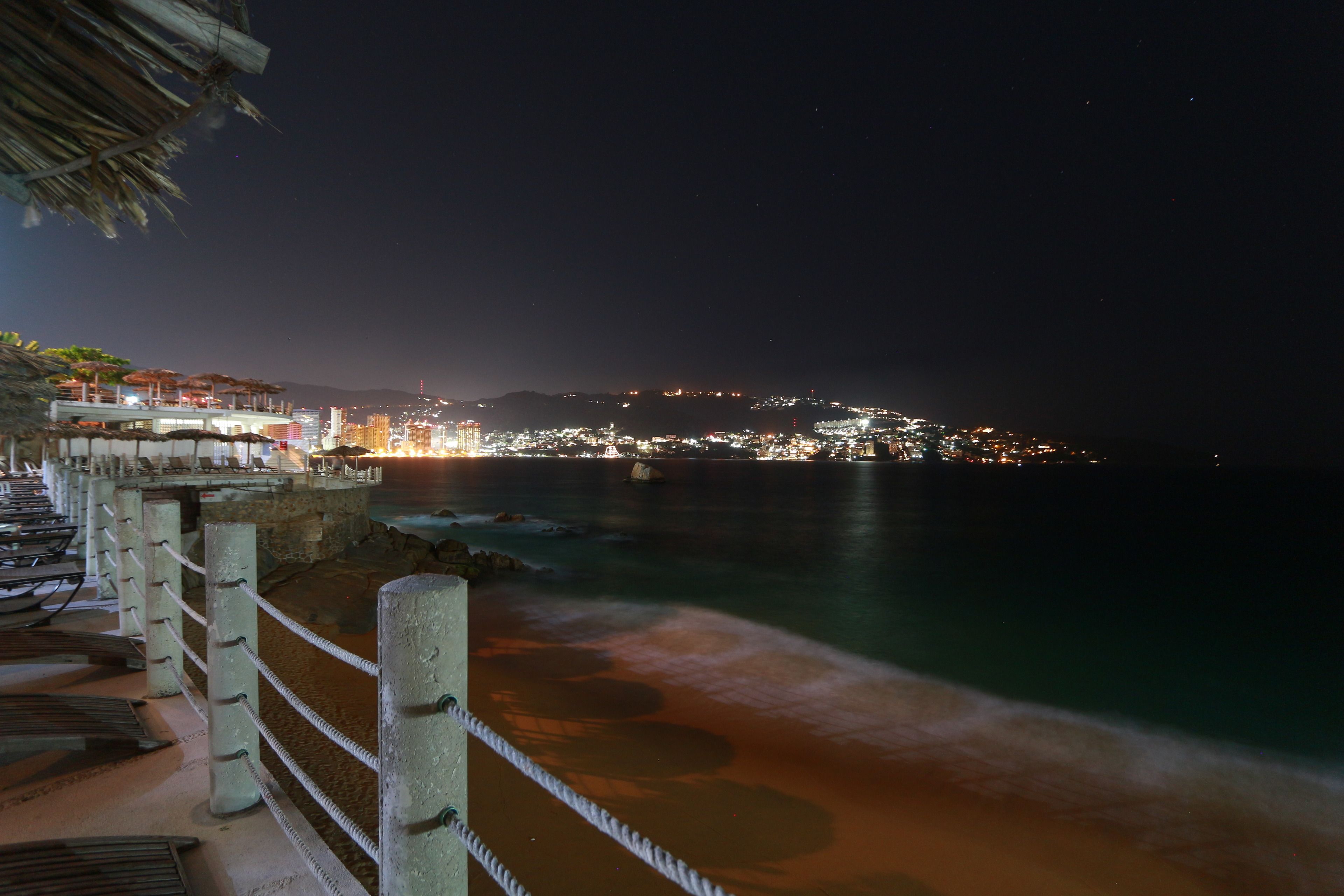 Un hotel costero con balcón que da al mar, una barandilla de cuerda y un paisaje urbano al fondo.