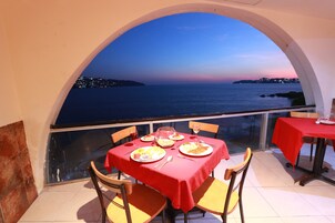 Mesa de jantar posta para dois, com toalha vermelha, posicionada sob uma grande janela em arco com vista para a costa.