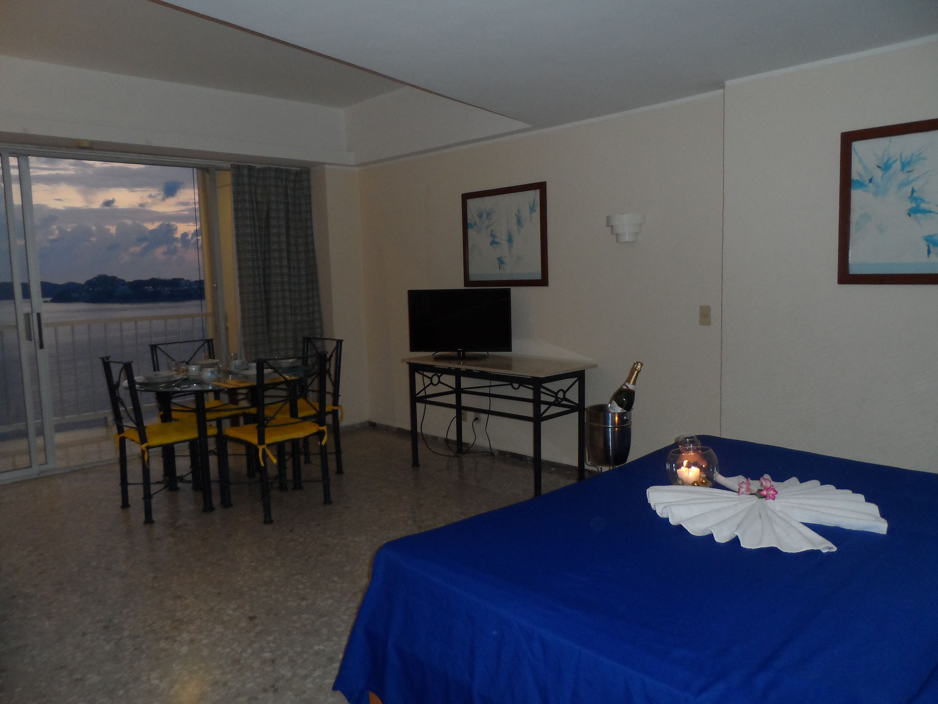 Habitación de hotel con balcón, una cama con cubierta azul, una mesa con sillas, un televisor y una botella de champán.