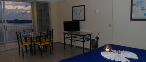 Habitación de hotel con balcón, una cama con cubierta azul, una mesa con sillas, un televisor y una botella de champán.