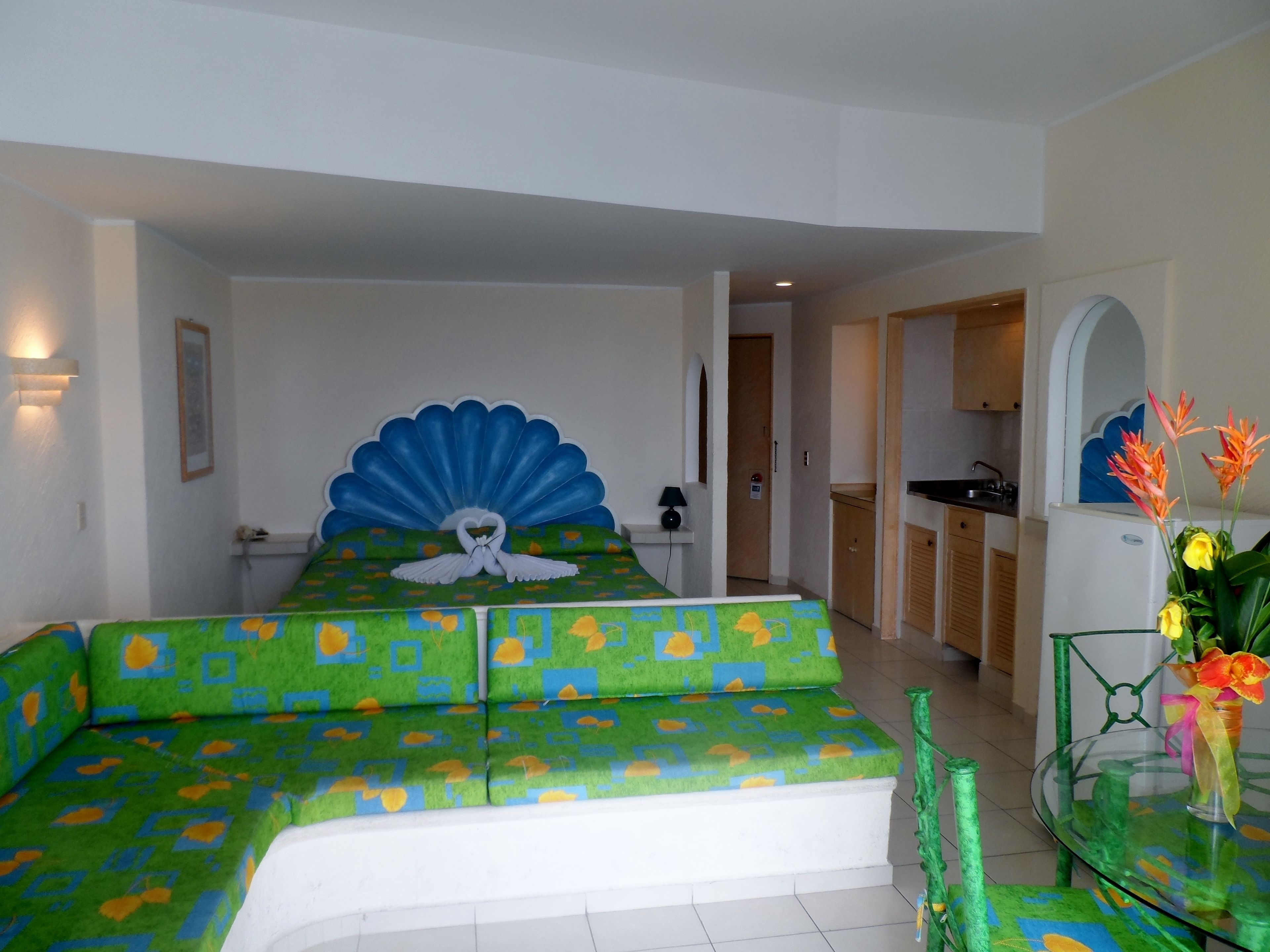 A hotel room with a bed featuring a blue scalloped headboard, a green and blue patterned sofa, and a glass table with a vase of flowers.