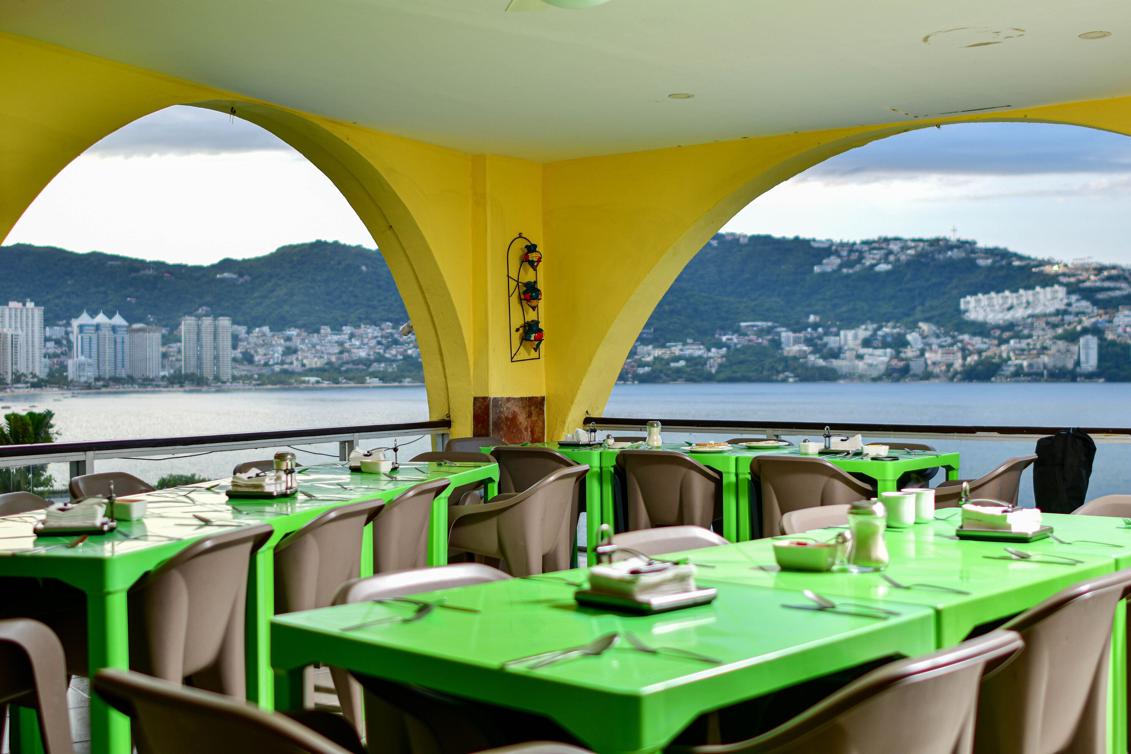 Un restaurante con mesas y sillas verdes, y una vista de la ciudad y el agua al fondo.