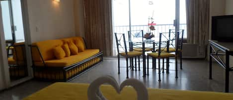 Habitación de hotel con cama, sofá amarillo, mesa de comedor con sillas, televisor y balcón con vistas.