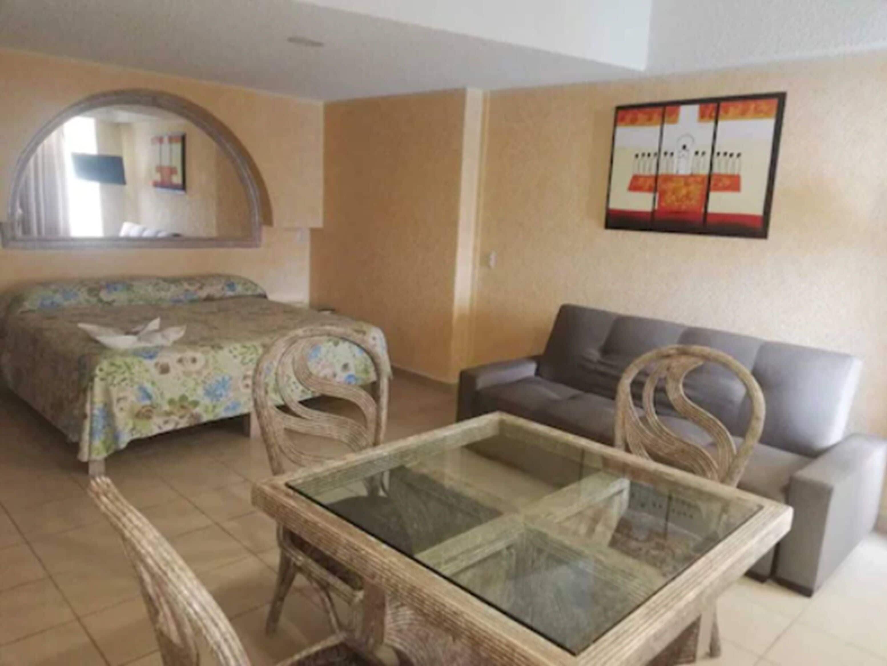 A room with a bed, a sofa, a glass-top table, wicker chairs, and a framed picture on the wall.