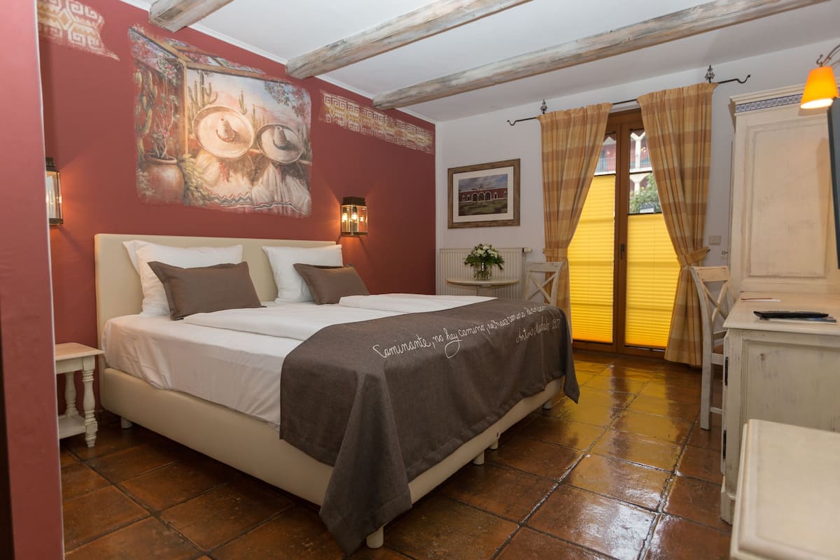 A bedroom with a bed, bedside tables, a desk, a chair, a lamp, a painting on the wall, and a window with curtains.