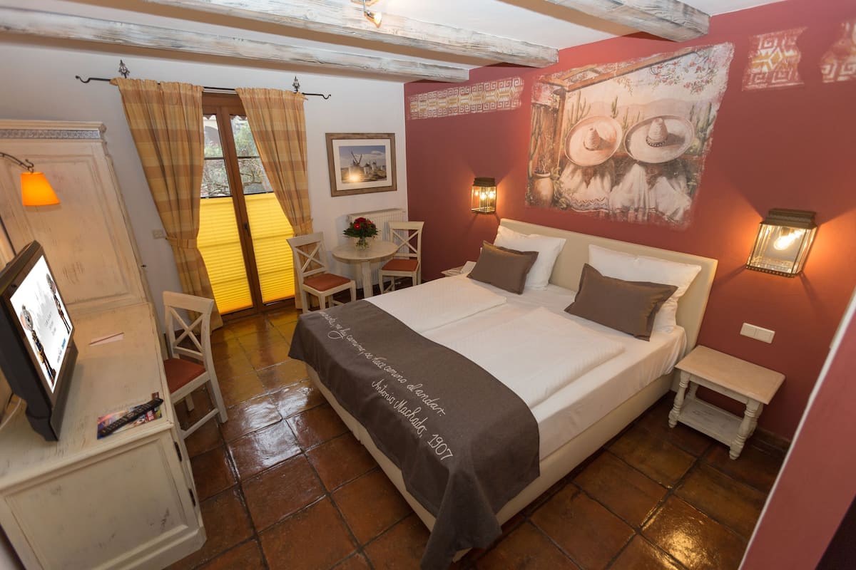 A bedroom with a bed, bedside tables, a desk with a chair, a small table with a vase, and a painting on the wall.