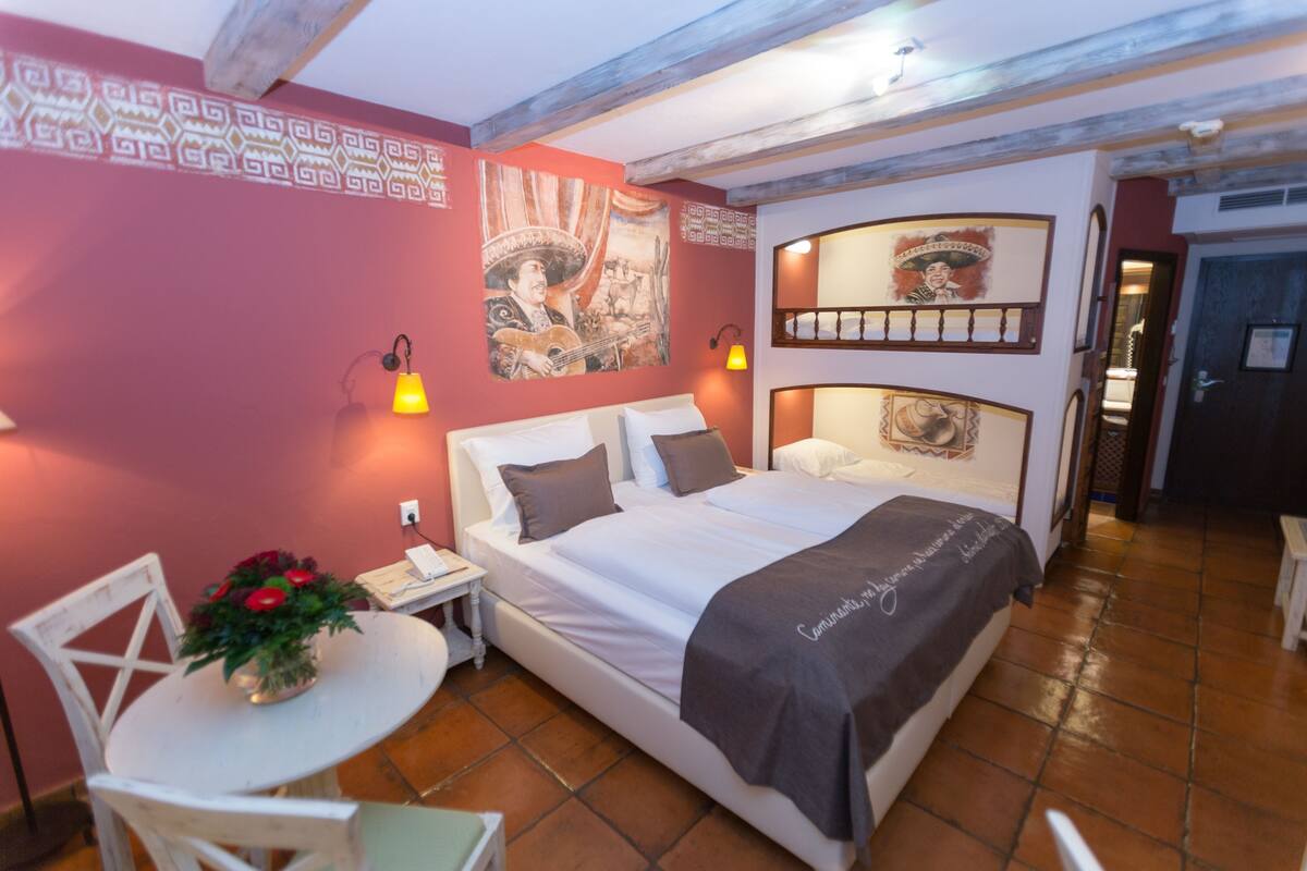 A bedroom with a bed, a small table with a plant, a chair, and a wall with decorative patterns and artwork.