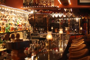 En bar med hyllor fulla av spritflaskor, en bardisk med barstolar och diverse barprylar.