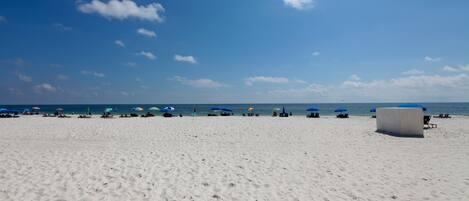 En bred sandstrand med en klarblå himmel och några få moln. Längs stranden finns flera blå parasoller och solstolar.