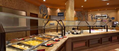 Uma área de buffet com várias estações de comida, uma placa indicando 'Aveia' e um pilar grande no centro.