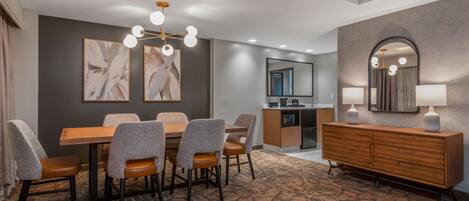 Une chambre d’hôtel dotée d’un coin repas comprenant une table et des chaises en bois, un lustre moderne, un grand miroir et un mini-bar.