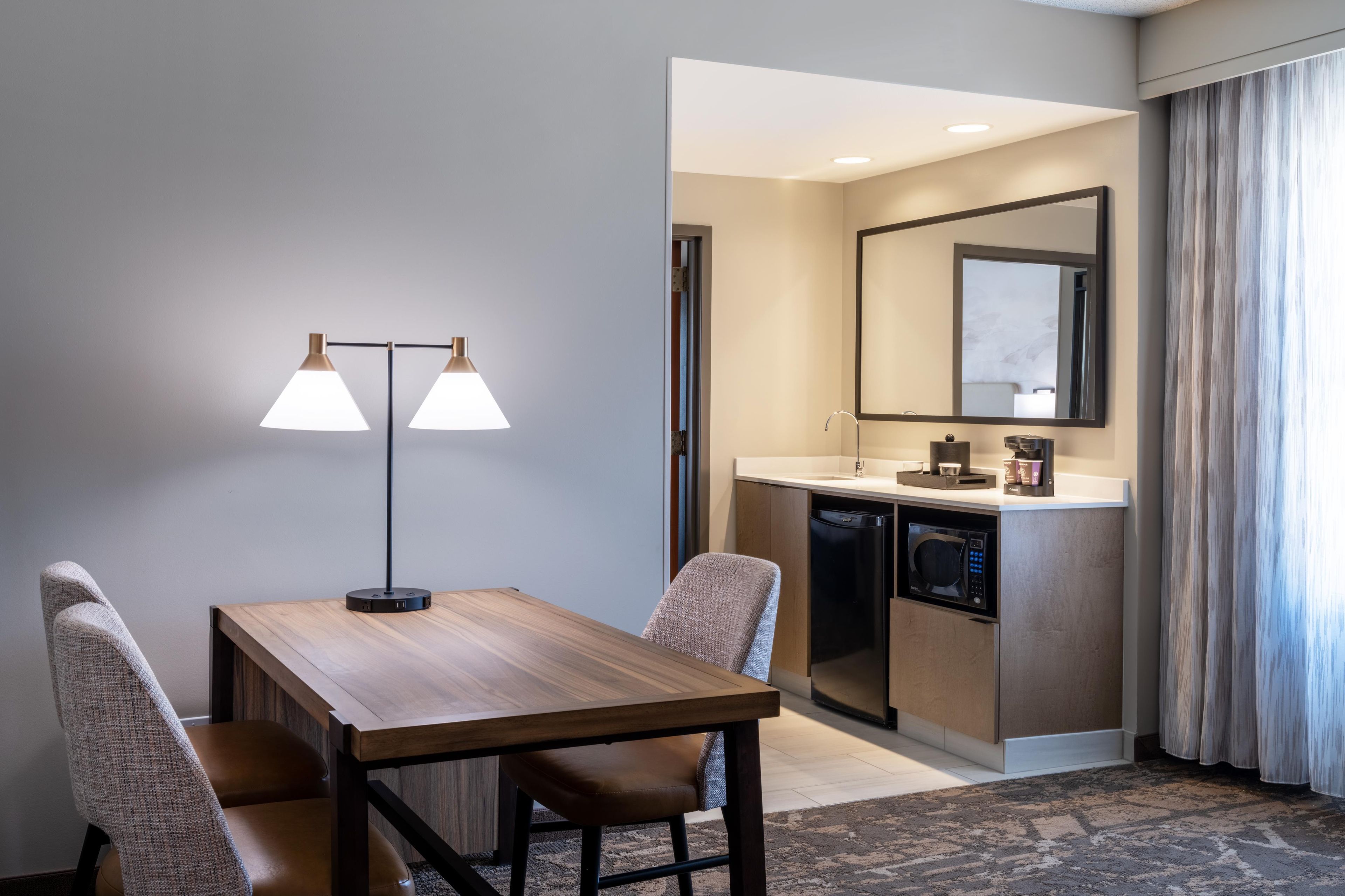 A hotel room with a wooden dining table, two chairs, a modern lamp, a kitchen area with a microwave, and a large mirror.