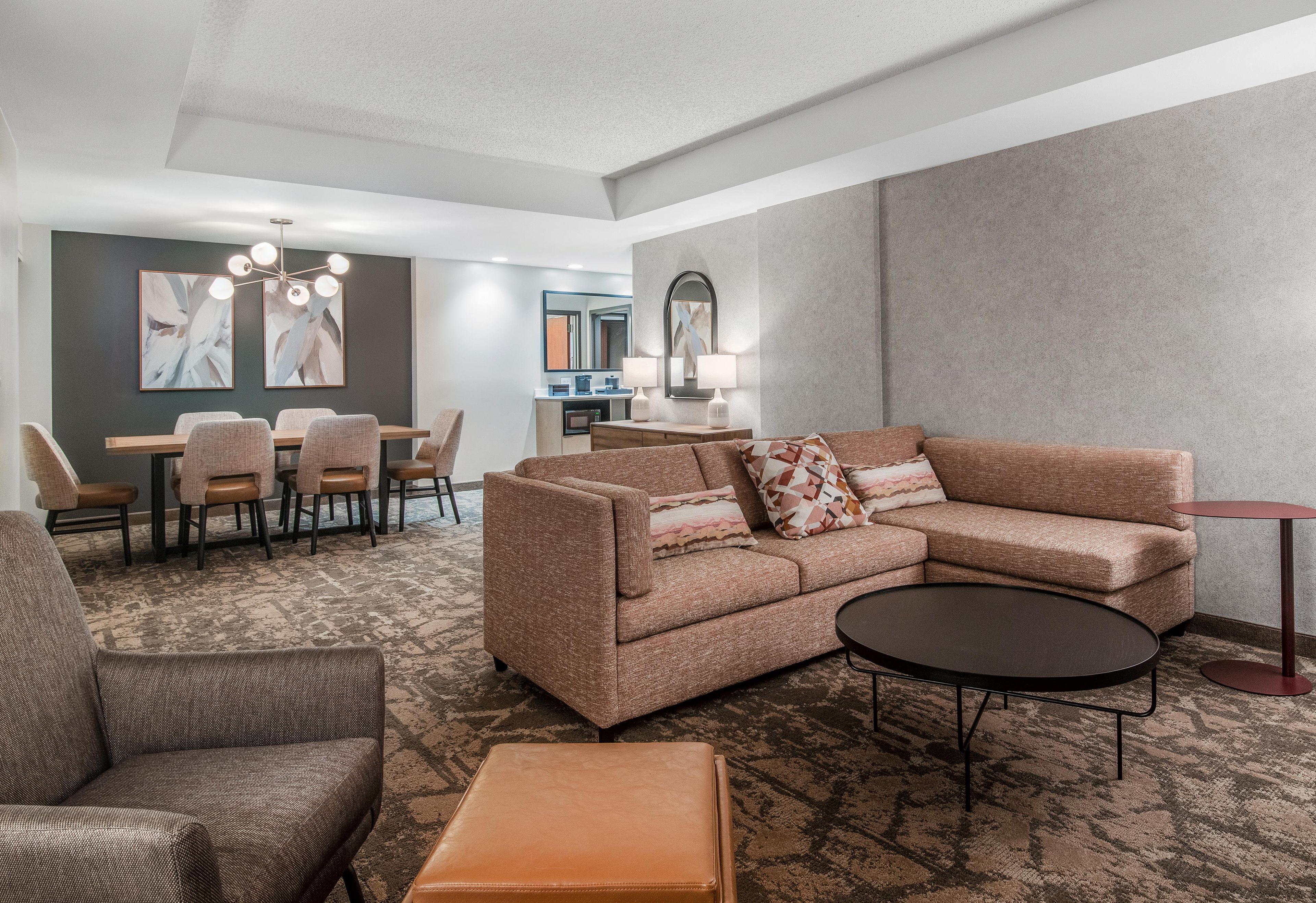 A modern hotel room with a sectional sofa, a small round table, a dining area with chairs, and a kitchenette.