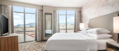 Quarto de hotel com cama grande, televisão e vista para o horizonte urbano através de amplas janelas.