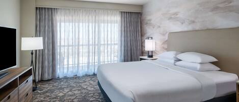 A hotel room with a large bed, a flat-screen TV, a desk with a lamp, and a view through sheer curtains.