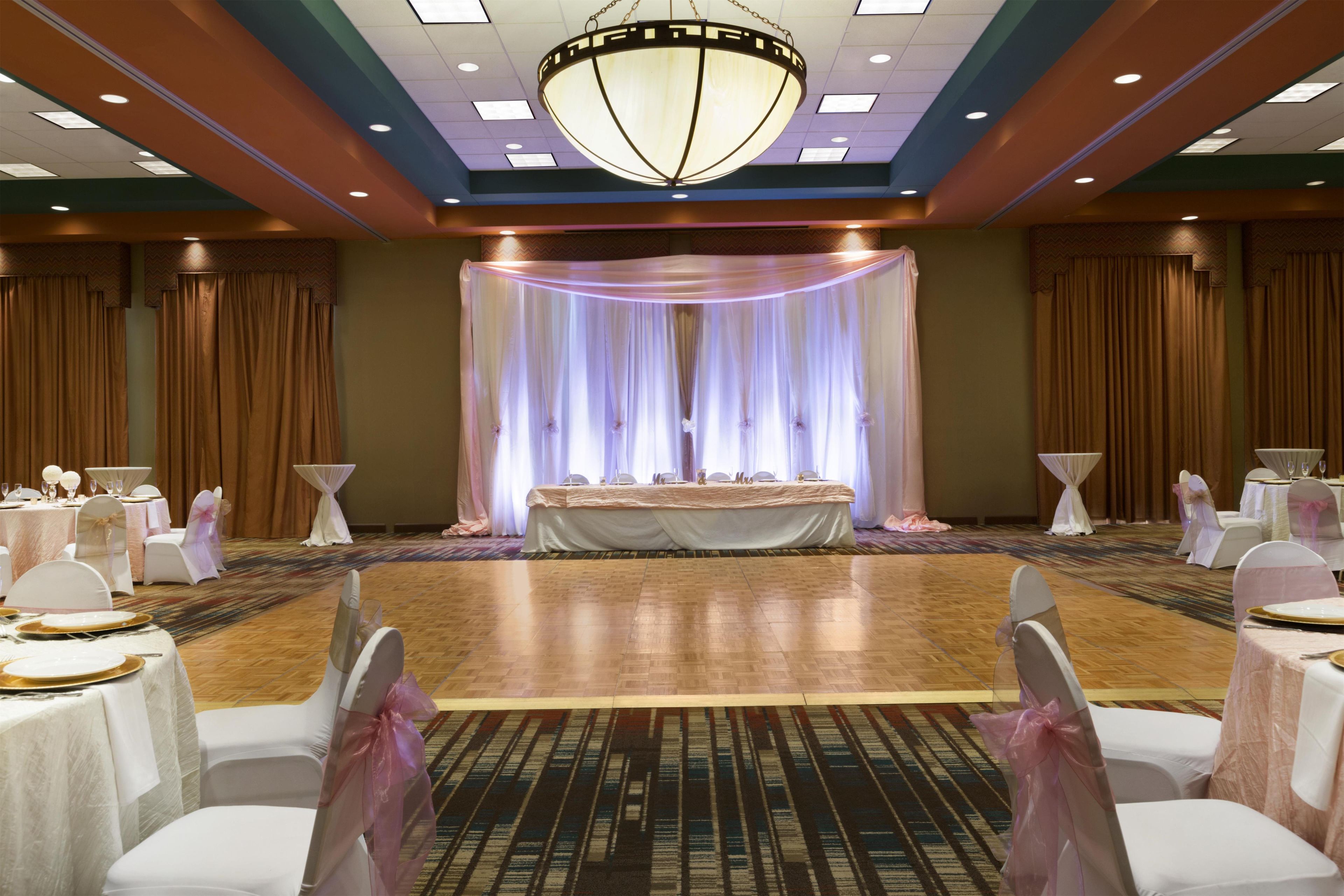 A banquet hall with a central stage, round tables with white tablecloths, and chairs with pink sashes.