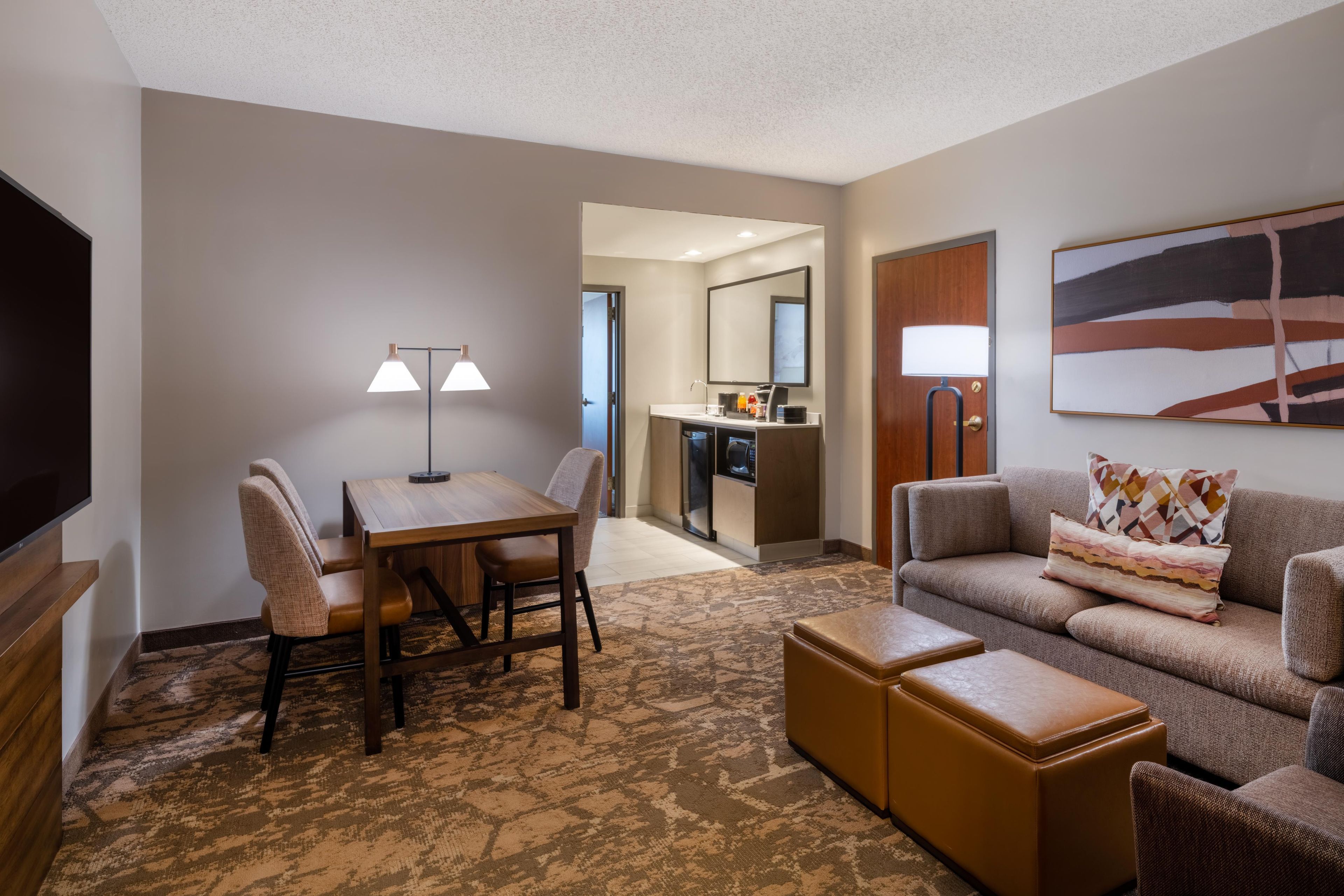 A hotel room with a dining table, two chairs, a sofa, a TV, a kitchen area, and a painting on the wall.
