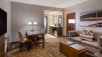 A hotel room with a dining table, two chairs, a sofa, a TV, a kitchen area, and a painting on the wall.